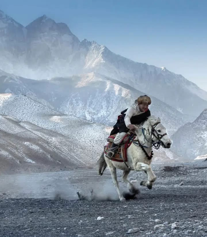A rider in a fur hat galloping a white horse across a stony valley below snow-covered peaks